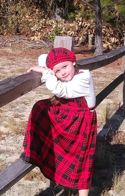 Alexis is registered to the contest to win money with this photo: dress, eye, face, grass, hat, headwear, human_body, leaf, magenta, pattern, people, person, plaid, plant, red, shawl, sitting, tartan, toddler, tree