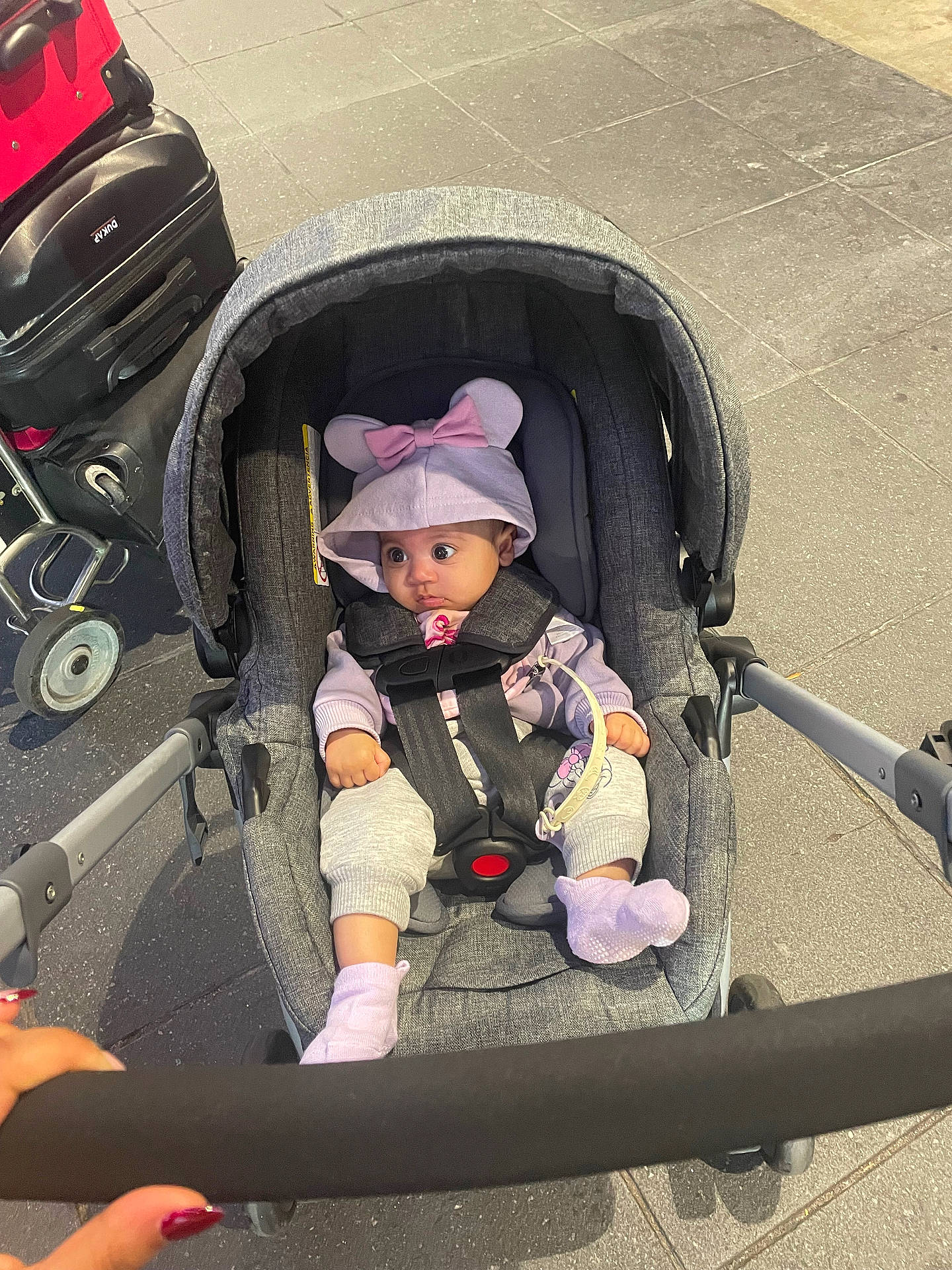 baby, baby_clothing, bow, car_seat, casual_clothing, child, curious, floor, hand, hoodie, infant, luggage, mouse_ears, person, purple, seat_belt, socks, stroller, tiled_floor, travel