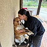 person, dog, chair, kiss, affection, indoor, outdoor, wall, window, floor, tile, casual_clothing, black_sweatshirt, brown_dog, white_fur, hand_holding, pet, animal, home, love