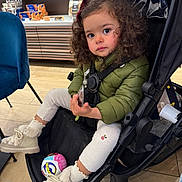 Liv a rejoint le concours — aidez-le/la à gagner de superbes lots ! toddler, child, stroller, curly_hair, green_jacket, boots, toy_ball, store, shelf, packaged_goods, floor, chair, indoor, warm_clothing, person, expression, seated, shopping_area, cute
