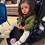 toddler, child, stroller, curly_hair, green_jacket, boots, toy_ball, store, shelf, packaged_goods, floor, chair, indoor, warm_clothing, person, expression, seated, shopping_area, cute