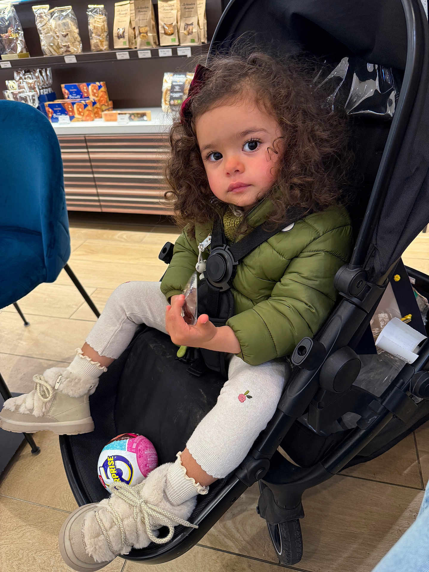 Liv a rejoint le concours — aidez-le/la à gagner de superbes lots ! toddler, child, stroller, curly_hair, green_jacket, boots, toy_ball, store, shelf, packaged_goods, floor, chair, indoor, warm_clothing, person, expression, seated, shopping_area, cute
