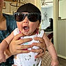 August is registered to the contest to win money with this photo: baby, sunglasses, onesie, infant, indoor, person, hand, cap, expression, furniture, mirror, ceiling_fan, animal_print, clothing, child, adult_hand, surprised, seated, home, cushion