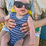 baby, child, sunglasses, tongue_out, striped_clothing, person_holding, sunlight, outdoor, grass, shorts, tshirt, reflection, hands, legs, casual, summer, seat, playful, fun, cute