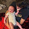 baby, child, couch, blanket, red_shorts, grey_onesie, smile, happy, indoor, person, holding, affection, family, hair, face, arm, seated, clothing, comfort, home