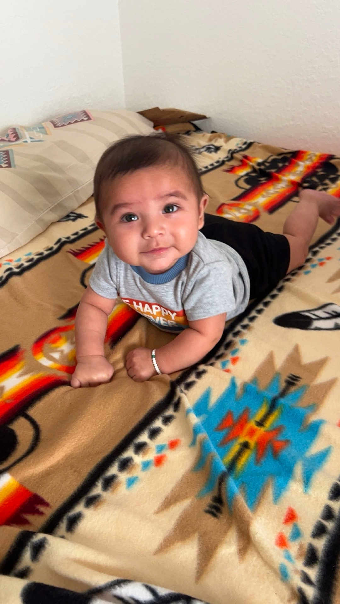 Dakota is registered to the contest to win money with this photo: baby, infant, child, blanket, patterned_blanket, toddler, indoor, bedroom, pillow, shorts, bracelet, smiling, face, cute, young_child, lying_down, looking_up, grey_shirt, skin, person