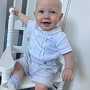 Hunter is registered to the contest to win money with this photo: baby, child, smiling, blue_eyes, white_shirt, checkered_shorts, sitting, chair, indoor, wooden_chair, happy, cute, footwear, sock, hand, face, portrait, infant, clothing, floor