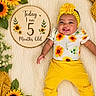 age_marker, baby, blanket, bright_colors, clothing, cute, decor, flower, greenery, happy, headband, infant, lying_down, portrait, smiling, socks, soft_texture, sunflower, wooden_plaque, yellow