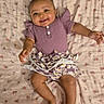 baby, child, smiling, purple_clothing, floral_skirt, blanket, patterned_blanket, lying_down, happy, cute, infant, human, person, indoors, skin, face, hand, foot, head, earrings