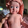 baby, infant, child, portrait, close_up, wide_eyes, smiling, head_wrap, pink, towel, bare_chested, held, adult_hand, indoor, bedroom, soft_lighting, bokeh, sitting, human_face, photography