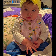 Isabella is registered to the contest to win money with this photo: baby, infant, face, headband, yellow_headband, dress, buttons, hands, bed, pillow, blanket, bedding, room, portrait, cute, sitting, eyes, cheeks, indoors, child