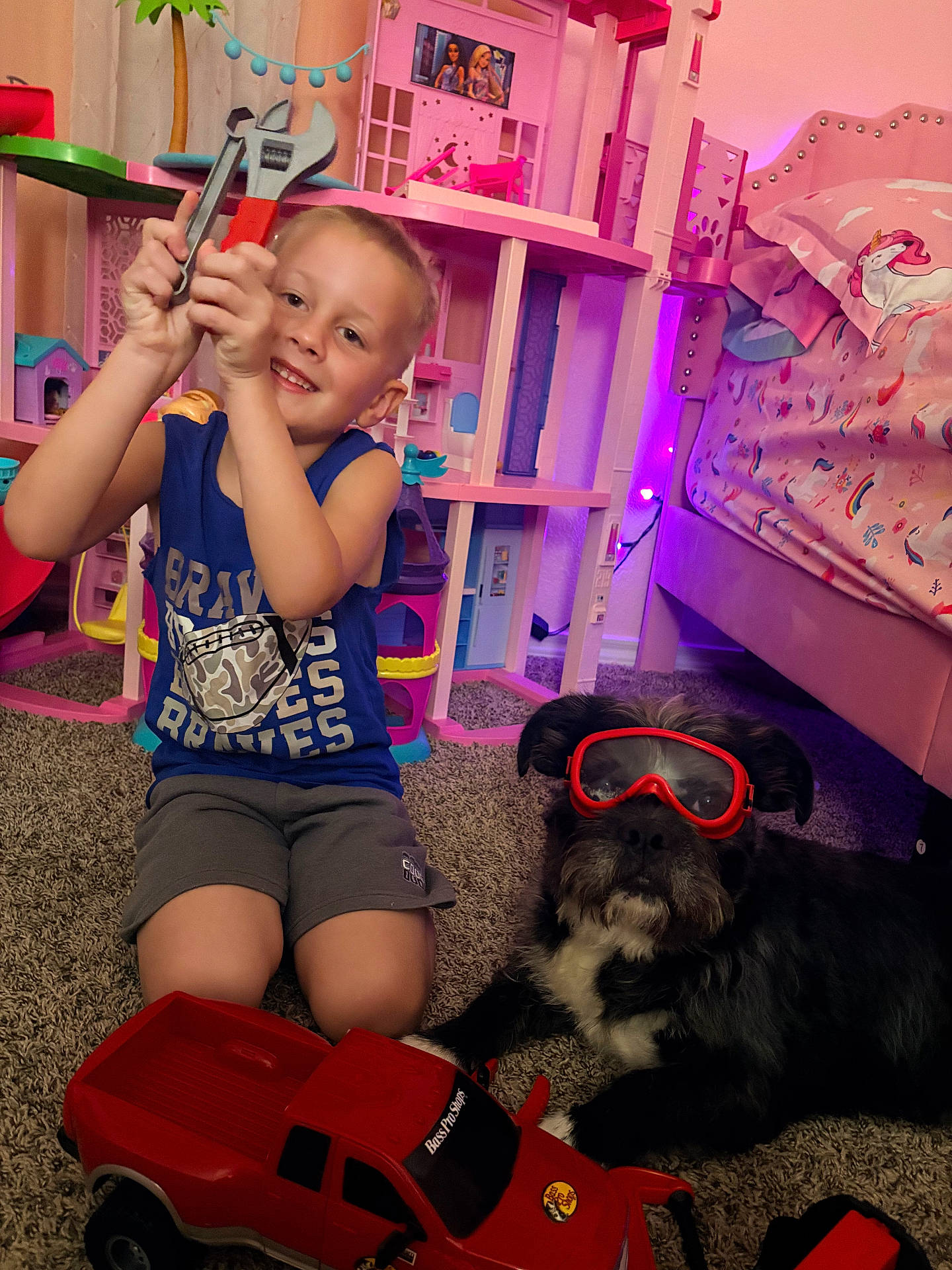 Sam is registered to the contest to win money with this photo: bed, bedroom, boy, carpet, child, dog, dollhouse, floor, furniture, goggles, happy, indoor, kid, pet, pink, playroom, smiling, toy_tools, toy_truck, unicorn_bedding