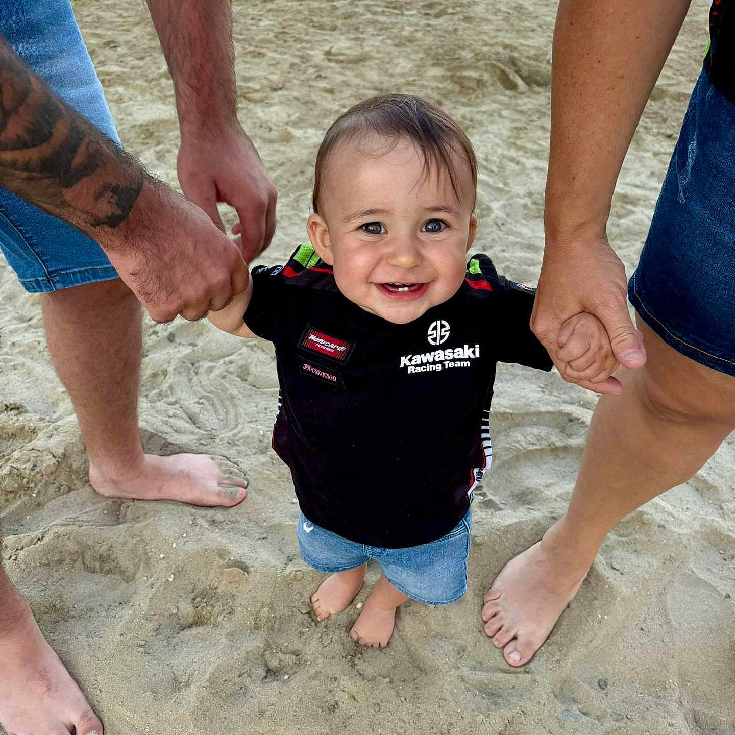 Tyméo participe au concours pour gagner de l'argent avec cette photo : adult, barefoot, beach, black_shirt, casual_clothing, child, denim_shorts, family, footwear_none, happy, holding_hands, outdoor, person, playing, sand, smile, summer, sunlight, tattoo, toddler