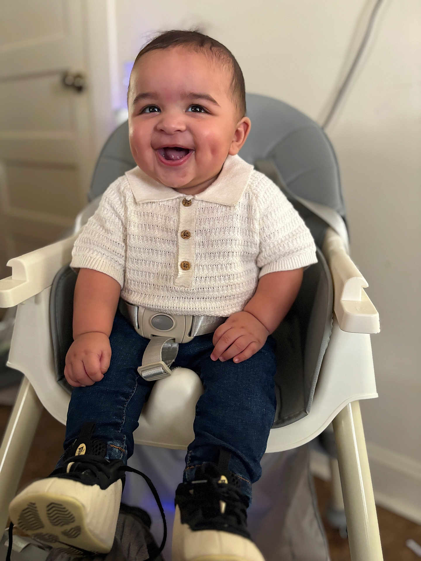 Trevell joined the competition — help win amazing prizes! baby, child, high_chair, smiling, dimples, white_shirt, knitwear, blue_jeans, sneakers, indoor, happy, seatbelt, cute, portrait, young_child, furniture, person, hands, eyes, face