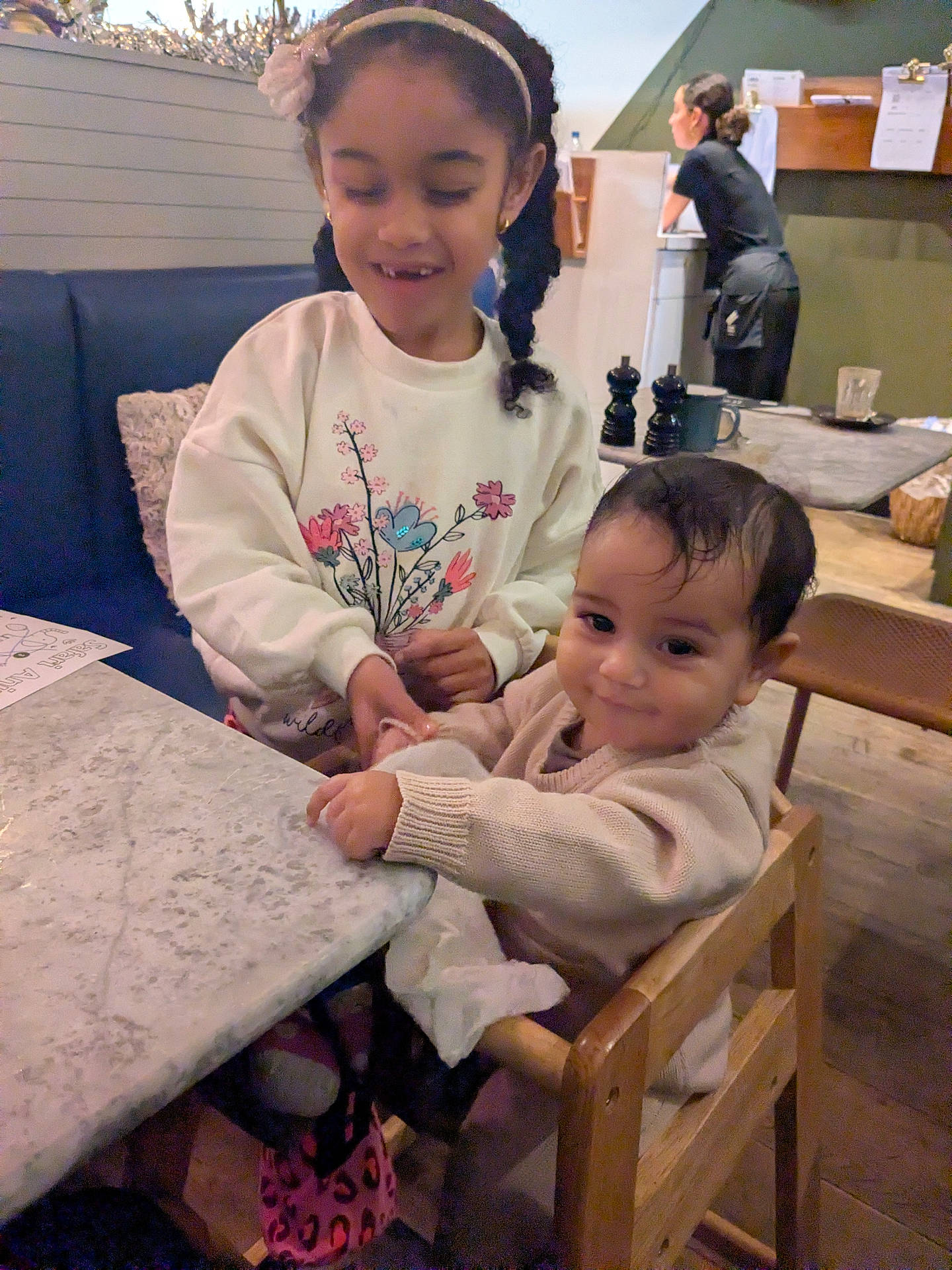 baby, background, cafe, casual, child, family, girl, hairband, happy, high_chair, indoor, person, restaurant, smile, sweater, table, toddler, waitress, woman, wooden_chair