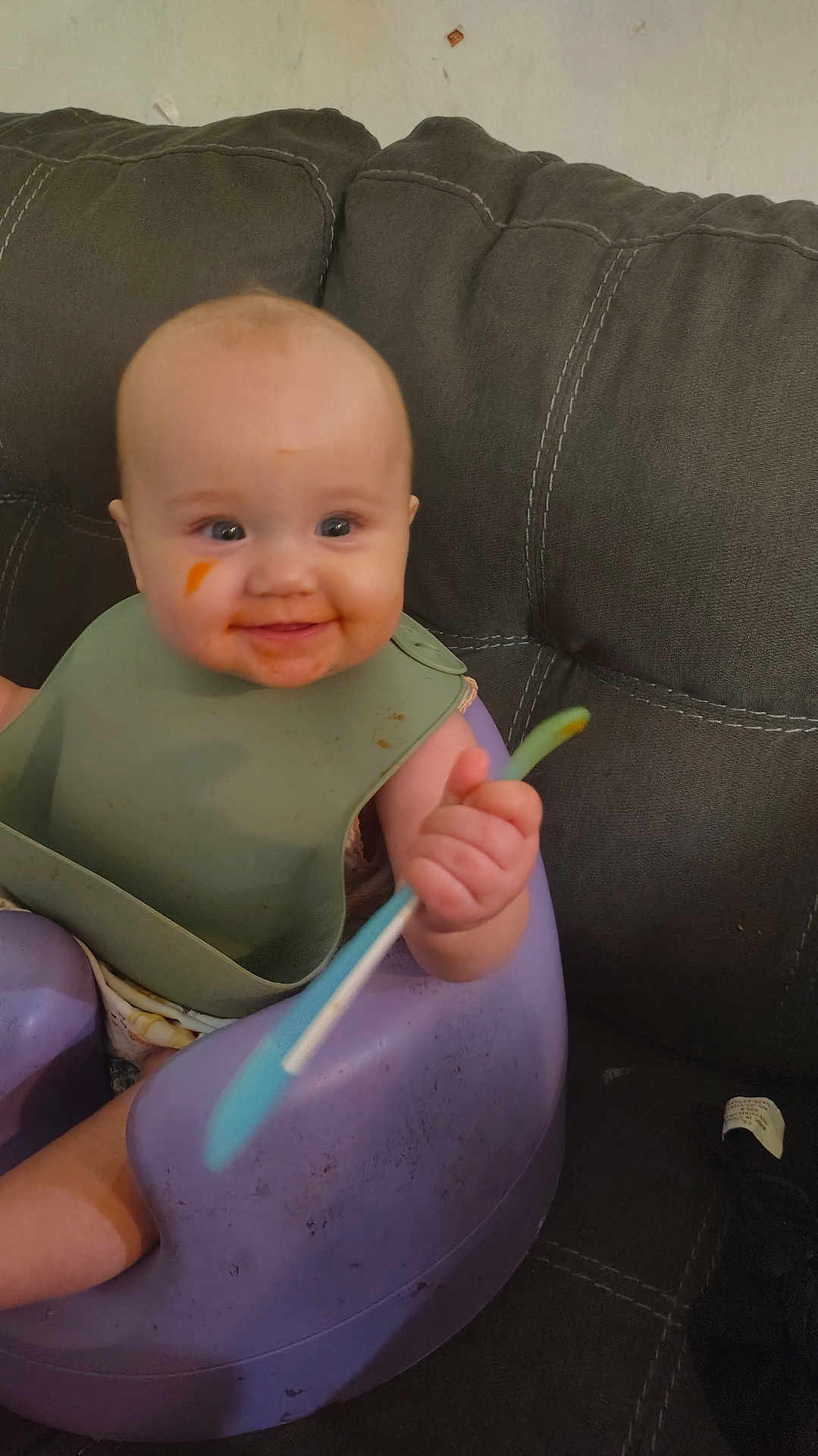 Isla is registered to the contest to win money with this photo: baby, smiling, bib, spoon, purple_baby_seat, couch, food_smear, infant, child, happy, indoor, person, seat, clothing, face, hand, feeding, messy, cute, young_child