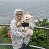 Harley is registered to the contest to win money with this photo: person, dog, hoodie, glasses, outdoor, ocean, coast, railing, greenery, waves, smile, pet, fluffy_dog, nature, casual_clothing, landscape, cloudy_sky, holding, leash, scenic