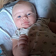 Riven is registered to the contest to win money with this photo: baby, infant, blue_eyes, blanket, onesie, toy, colorful, hand, face, skin, cute, indoor, child, person, furniture, wood, soft, expression, lying_down, closeup