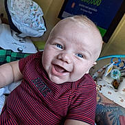 Riven is registered to the contest to win money with this photo: baby, smiling, blue_eyes, red_shirt, striped_clothing, tattoo, arm, baby_seat, television, indoor, happy, person, child, closeup, infant, skin, face, mouth_open, cheeks, cute