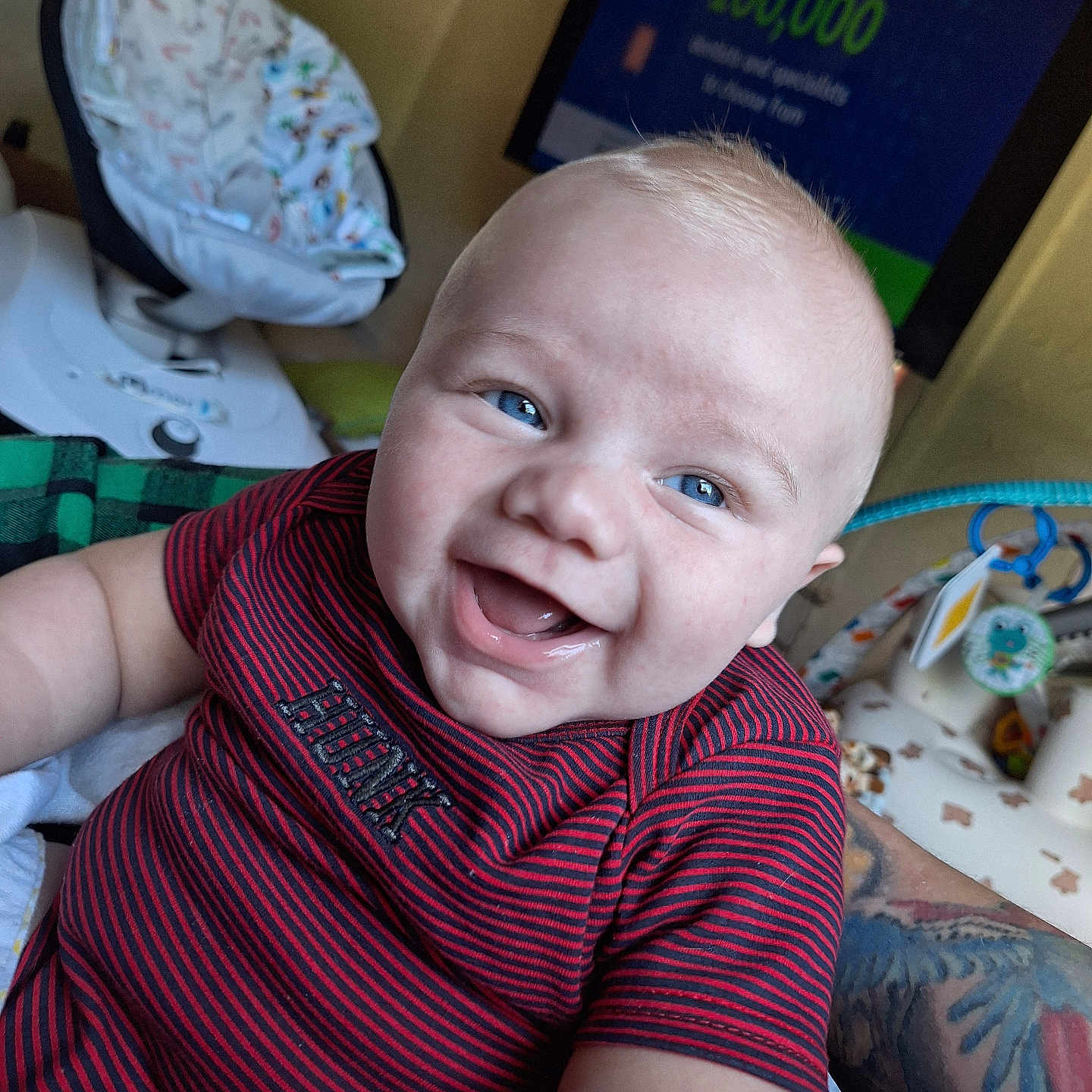 Riven is registered to the contest to win money with this photo: arm, baby, baby_seat, blue_eyes, cheeks, child, closeup, cute, face, happy, indoor, infant, mouth_open, person, red_shirt, skin, smiling, striped_clothing, tattoo, television