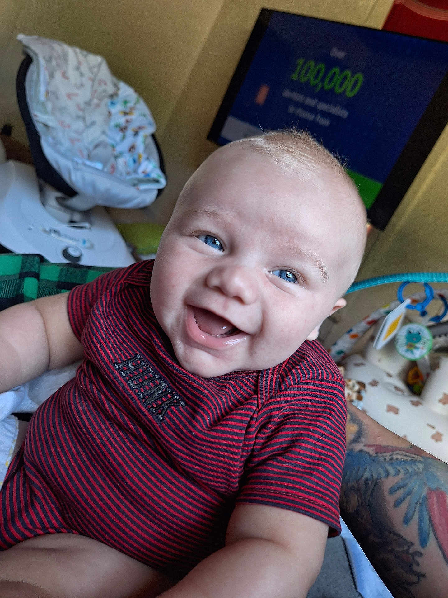 Riven is registered to the contest to win money with this photo: baby, smiling, blue_eyes, red_shirt, striped_clothing, tattoo, arm, baby_seat, television, indoor, happy, person, child, closeup, infant, skin, face, mouth_open, cheeks, cute