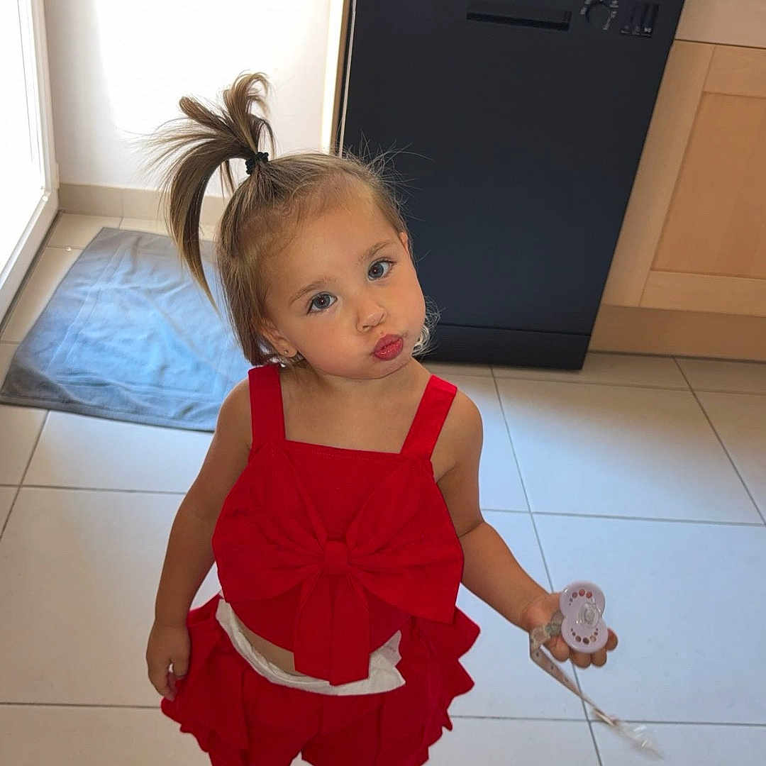 Miléna a rejoint le concours — aidez-le/la à gagner de superbes lots ! bow, cabinet, child, cute, dishwasher, expression, floor_tiles, girl, hair, indoors, kitchen, light, pacifier, playful, ponytail, pouty_face, red_clothing, sandals, standing, toy