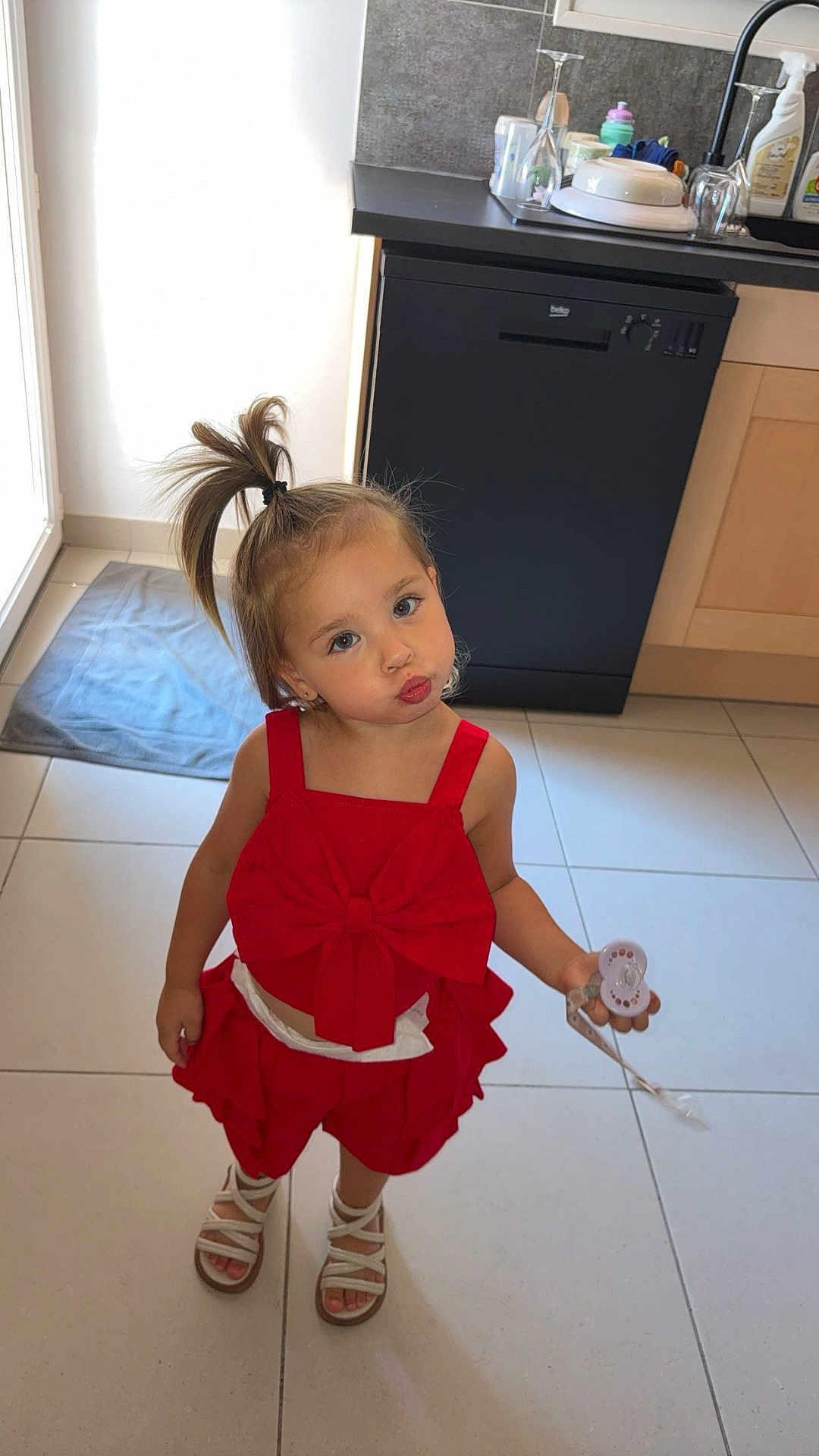 Miléna a rejoint le concours — aidez-le/la à gagner de superbes lots ! child, girl, red_clothing, bow, ponytail, pacifier, sandals, kitchen, floor_tiles, dishwasher, cabinet, playful, pouty_face, standing, indoors, light, expression, hair, toy, cute