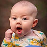 baby, infant, child, face, expression, mouth_open, clothing, floral_pattern, person, shoulder, holding, cute, portrait, skin, head, eyes, hand, outdoor, blurry_background, young