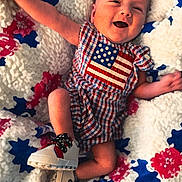 Knox is registered to the contest to win money with this photo: baby, infant, smiling, plaid_clothing, american_flag, blanket, knitted_blanket, red, white, blue, sneakers, feet, face, happy, cute, laying_down, child, patterned_blanket, cozy, apparel