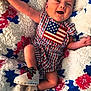 baby, infant, smiling, plaid_clothing, american_flag, blanket, knitted_blanket, red, white, blue, sneakers, feet, face, happy, cute, laying_down, child, patterned_blanket, cozy, apparel