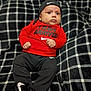 Eli Alexander is registered to the contest to win money with this photo: baby, infant, clothing, nike, hat, red_shirt, black_pants, blanket, plaid, pattern, footwear, portrait, indoor, person, cute, fashion, sitting, expression, child, young