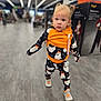 aisle, baby, black, child, clothing, curious, cute, floor, ghost_pattern, hair, indoors, orange, pacifier, person, portrait, shoes, store, toddler, walking, young