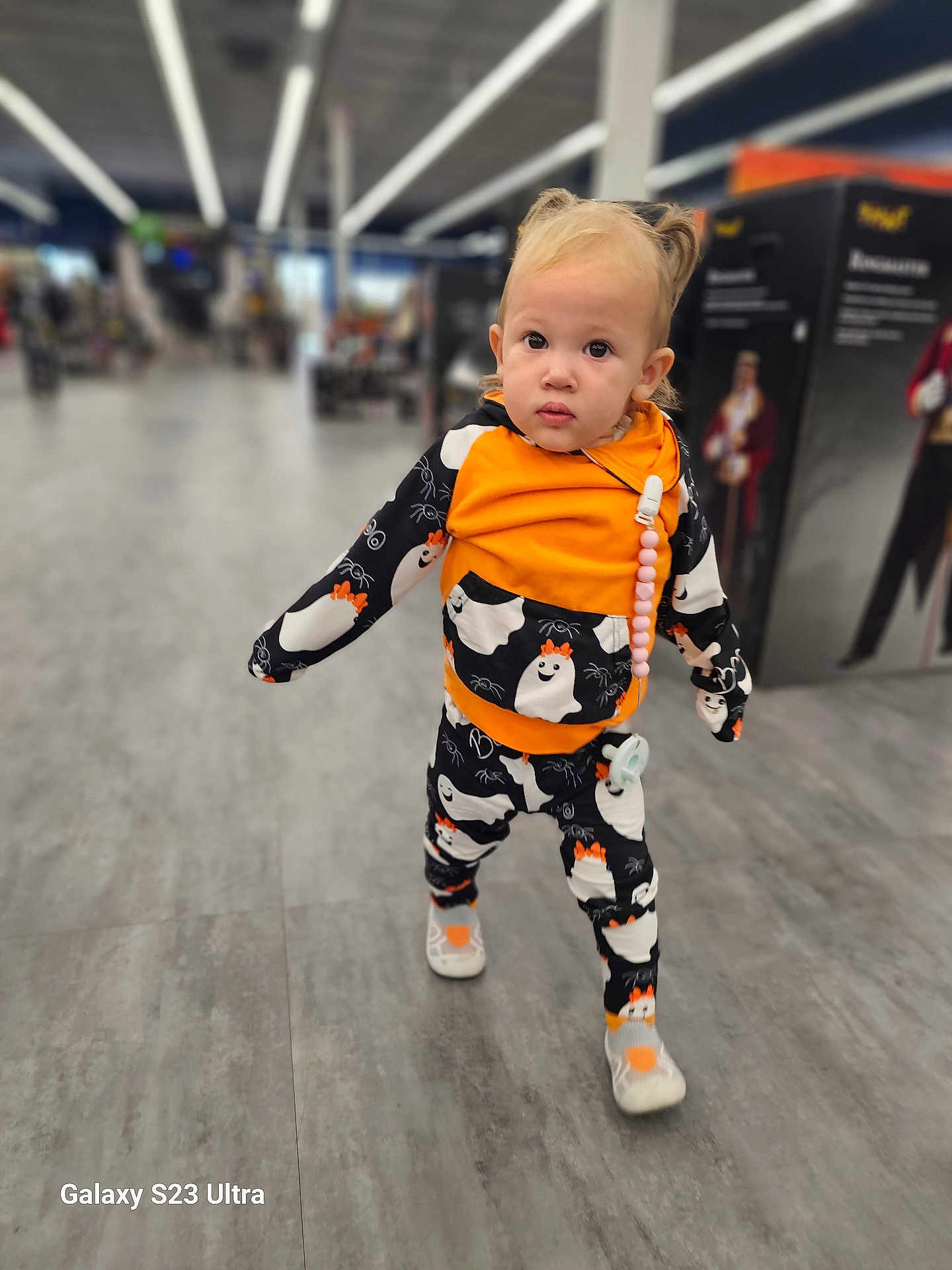 Xayna is registered to the contest to win money with this photo: toddler, child, person, walking, clothing, orange, black, ghost_pattern, shoes, hair, indoors, store, aisle, floor, pacifier, curious, cute, young, portrait, baby