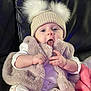 baby, child, hat, pom_poms, fluffy_vest, white_clothing, couch, blanket, tongue_out, cute, infant, indoor, person, sitting, cozy, playful, soft_texture, warm_clothing, pink_blanket, smiling