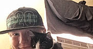 Oreo is registered to the contest to win money with this photo: person, cat, kitten, cap, indoor, selfie, black_and_white_cat, smile, face, hand, curtain, air_conditioner, wall, couch, light, clothing, pet, feline, cute, cozy