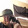 person, cat, kitten, cap, indoor, selfie, black_and_white_cat, smile, face, hand, curtain, air_conditioner, wall, couch, light, clothing, pet, feline, cute, cozy