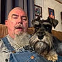 man, dog, schnauzer, beard, mustache, overalls, couch, indoor, portrait, pet, home, curtain, framed_photos, casual, blurred, relaxed, face, furniture, companion, seated