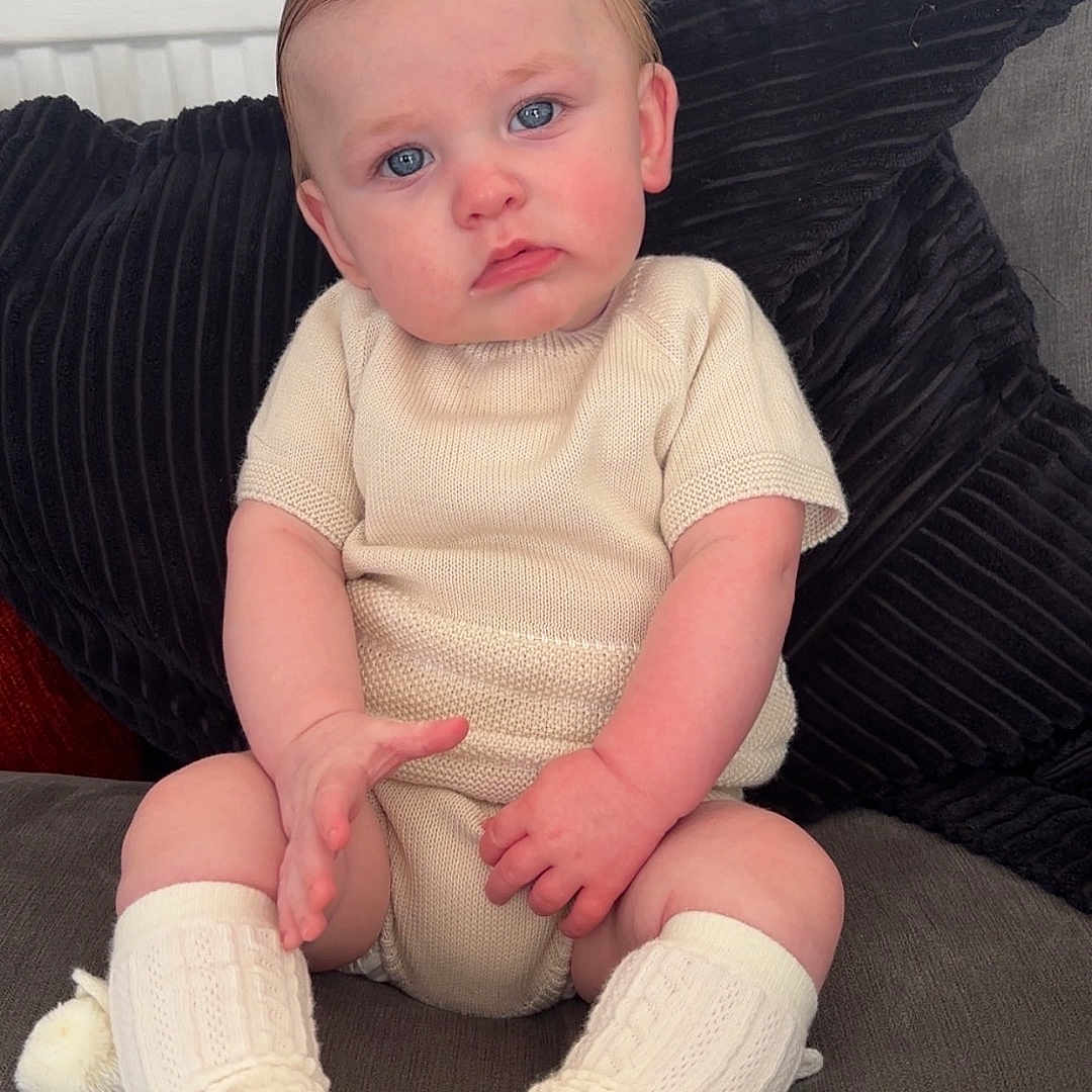 Vinnie is registered to the contest to win money with this photo: baby, beige_outfit, blue_eyes, child, comfort, couch, cozy, expression, footwear, hair, hand, indoor, infant, knitted_socks, pillow, portrait, seated, skin, soft_shoes, textile