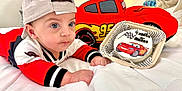 Matheo is registered to the contest to win money with this photo: baby, infant, child, cap, red_clothing, white_bedspread, plush_toy, toy_car, cake, celebration, portrait, indoors, cute, face, hands, blue_eyes, car_design, soft_toy, laying_down, hat