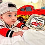 baby, infant, child, cap, red_clothing, white_bedspread, plush_toy, toy_car, cake, celebration, portrait, indoors, cute, face, hands, blue_eyes, car_design, soft_toy, laying_down, hat