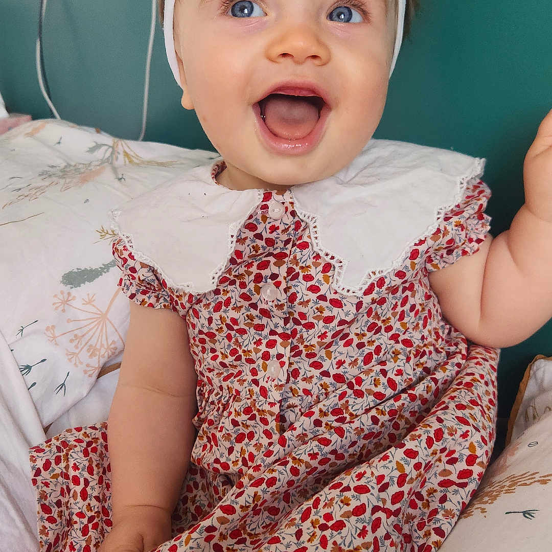 Giulia participe au concours pour gagner de l'argent avec cette photo : baby, bed, blue_eyes, cheerful, child, collar, cute, floral_dress, happy, headband, indoor, infant, person, pillow, portrait, sitting, skin, smiling, white_bow, young