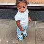 blue_bow, blue_sneakers, casual_clothing, child, curly_hair, curly_hair_bun, cute, earrings, fashion, floor_tiles, indoor, light_blue_jeans, person, portrait, reflection, standing, toddler, white_shirt, window, young_child