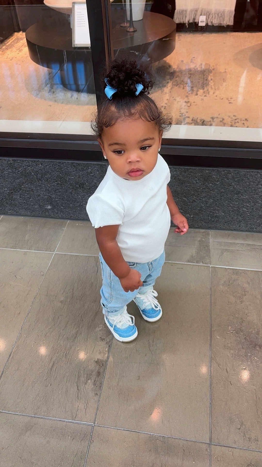 Rhemi is registered to the contest to win money with this photo: blue_bow, blue_sneakers, casual_clothing, child, curly_hair, curly_hair_bun, cute, earrings, fashion, floor_tiles, indoor, light_blue_jeans, person, portrait, reflection, standing, toddler, white_shirt, window, young_child