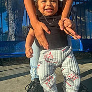 Aries is registered to the contest to win money with this photo: toddler, child, smiling, happy, outdoor, playground, adult, arms, lifting, portrait, face, black_shirt, patterned_pants, sneakers, blue_bench, sunlight, park, play, standing, family