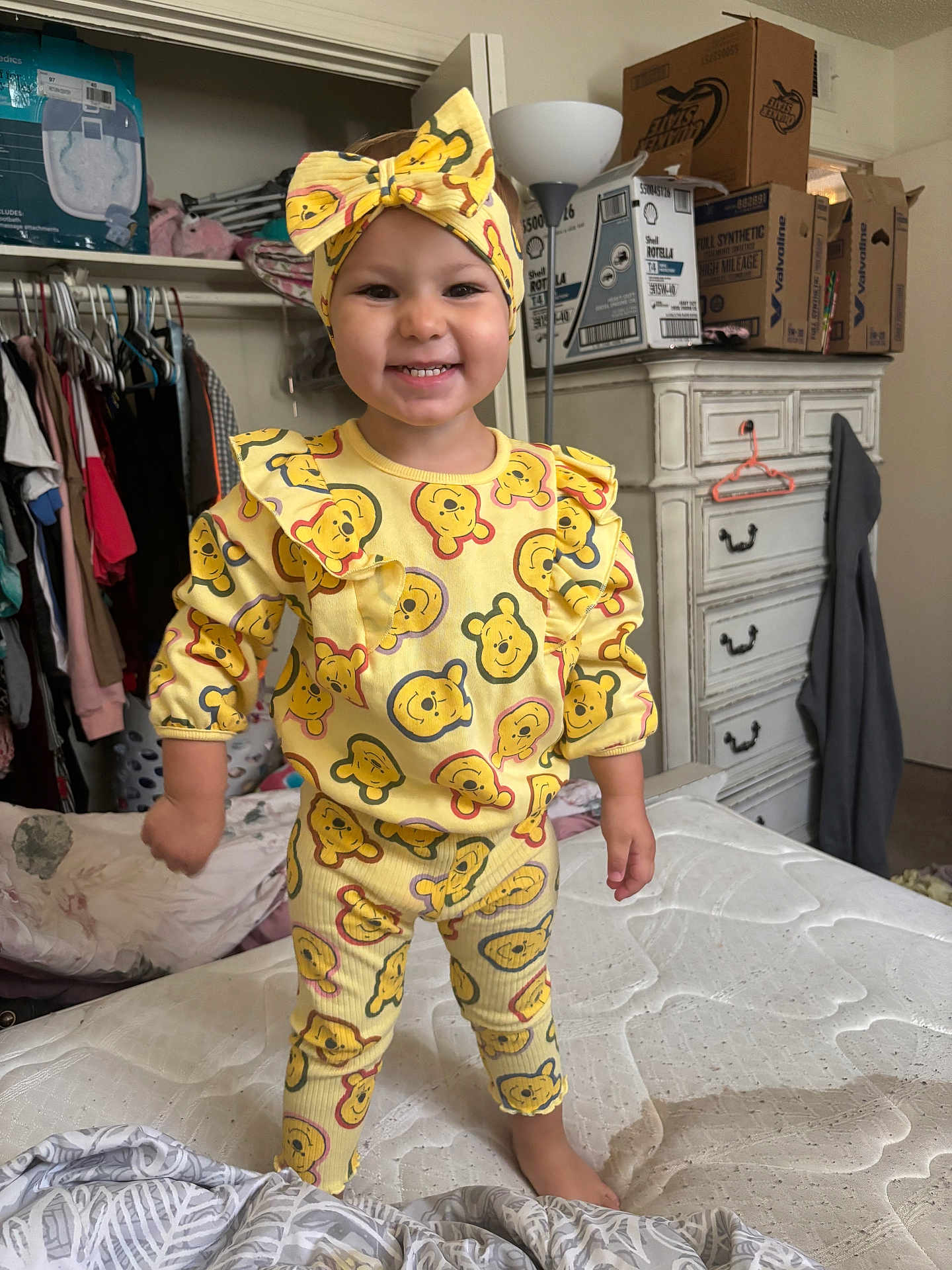 Athena joined the competition — help win amazing prizes! toddler, child, smiling, yellow_clothing, headband, bedroom, dresser, boxes, mattress, standing, indoors, clothing_pattern, cartoon_bear, happy, person, footwear_barefoot, furniture, hanger, lamp, closet