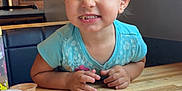 Athena joined the competition — help win amazing prizes! toddler, child, smiling, blue_bow, blue_shirt, wooden_table, food_container, restaurant, indoor, cute, happy, face, hands, seat, wall_art, poster, brick_wall, playful, eating, portrait