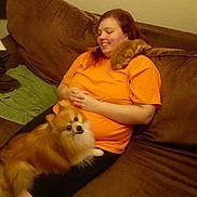 Aslain is registered to the contest to win money with this photo: brown_couch, casual, cat, comfortable, cozy, fluffy_dog, furniture, happy, home, indoor, lap, orange_shirt, pet, pomeranian_dog, relaxing, resting, shoulder, small_cat, smiling, woman