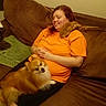 brown_couch, casual, cat, comfortable, cozy, fluffy_dog, furniture, happy, home, indoor, lap, orange_shirt, pet, pomeranian_dog, relaxing, resting, shoulder, small_cat, smiling, woman