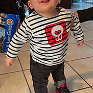 Tom participe au concours pour gagner de l'argent avec cette photo : toddler, child, indoor, striped_shirt, sneakers, red, black, jeans, floor_tiles, person, smiling, tongue_out, standing, hand_support, background, bag, chair, table, home, happy