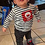 toddler, child, indoor, striped_shirt, sneakers, red, black, jeans, floor_tiles, person, smiling, tongue_out, standing, hand_support, background, bag, chair, table, home, happy